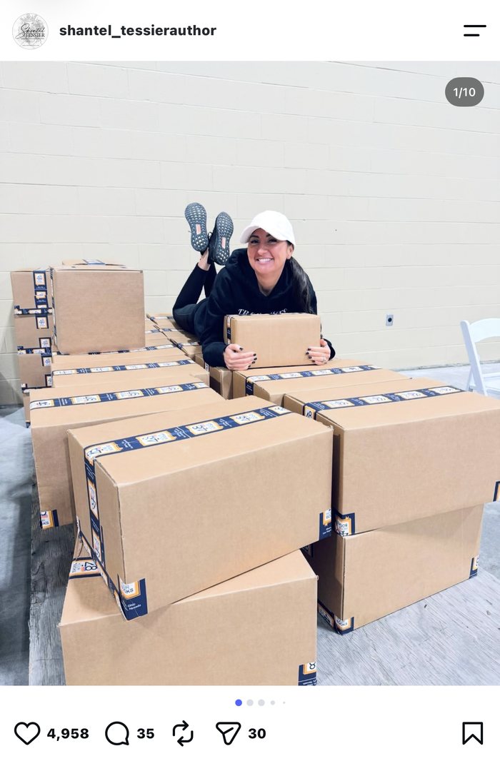 Author Shantel Tessier posing across stacked shipping boxes at the Sweetgrass 2024 signing event